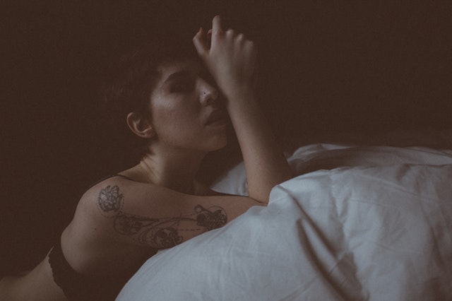 Žena v spodnej bielizni a výrazným tetovaním na ruke kľačí pri posteli