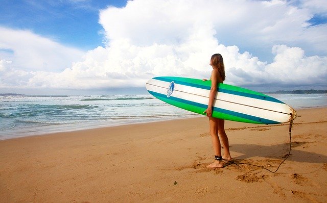 Žena v plavkách a so surfom v ruke stojí na pláži a pozerá na more.jpg